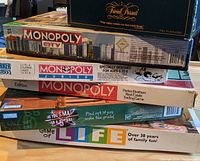 Stack of six board games including Trivial Pursuit, three types of Monopoly (original, City, Junior), Life, and Are You Smarter Than a 5th Grader.
