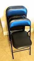 Four black folding chairs stacked together against a wall on tiled floor.