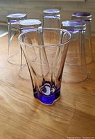 Six drinking glasses with purple tint, one standing upright in front and five inverted behind on a wooden surface.