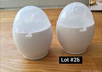 Two white egg cookers with clear plastic lids viewed from the front on a wooden surface.