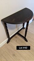 Photo of a dark stained halfmoon wooden table with three turned legs against a wall on a wooden floor.