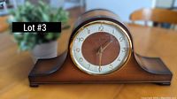 Freestanding wooden mantle clock with round dial, brass bezel, and dark wood base shown on wooden table with plant in background.