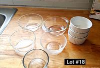 Photo of 3 white ceramic ramekins stacked beside 5 clear glass mini dishes on a wooden counter near a sink and tiled backsplash.
