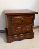 Wooden night table with three drawers shown against a white background. Visible details include decorative brass hardware and minor wear on the base.