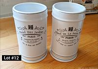 Two white ceramic cylindrical holders side by side on a wooden surface with Paris-themed printed text and designs on each.