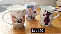 Three ceramic mugs placed on a wooden surface, brightly lit in natural light. Left mug with cats, center with floral and ladybug, right with purple cat design.