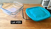 Two clear square glass baking dishes stacked on a wooden countertop with a turquoise plastic lid lying beside them marked 'PYREX'.