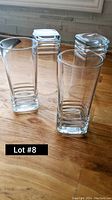 Photo of four clear tall glass tumblers with square bases, standing on a wooden surface showing their shape and condition.