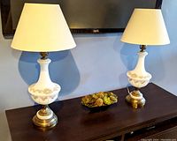 Pair of white glass table lamps with gold floral designs on metal bases, each with white fabric shades, placed on dark wood console table beneath a wall-mounted TV.