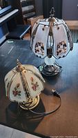 Two vintage touch lamps on dark wood surface, one smaller with cream rose painted shade and the other larger with floral painted panels and textured glass base