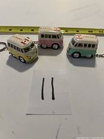 Three VW van key chains lined up on a flat surface with measuring tape behind them showing size. Yellow van on left, pink center, teal right.