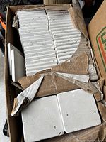 Box with stacked white ceramic tiles, some tiles visible on top, showing square shape and uniform white color.