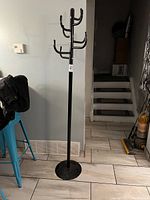 Full view of the tall black metal coat rack standing on tiled floor near a wall and steps.