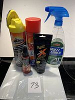 Photo showing six assorted car care products arranged in two rows against a white wall background. Includes yellow Armor All can, red aerosol can, black Meguiar's ScratchX tube, clear Turtle Wax spray bottle, and two smaller red bottles.