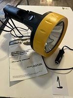 Photo of black and yellow Coleman Powermate collapsible spotlight with owner's manual and car power adapter plug visible.