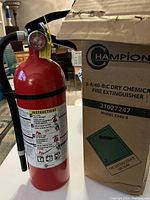 Front view of the red Champion dry chemical fire extinguisher next to its box showing label with instructions and brand details.