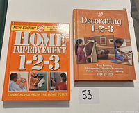 Photo showing two Home Depot books, 'Home Improvement 1-2-3' and 'Decorating 1-2-3' arranged side by side on a white surface with lot number card 53.