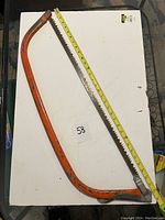 Photo of large pruning saw with orange handle frame and blade about 34 inches long on white background with yellow measuring tape visible.
