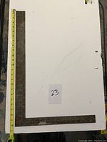 Large metal square ruler placed on a white surface with measuring tape showing length, marked with a paper labeled '23'.