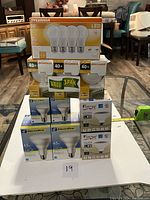 Stack of LED light bulb boxes from Sylvania, ElectriPro, Fortec brands including various wattages and types such as standard and flood bulbs.