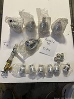 17-piece lot of new plumbing items including multiple chrome shower and faucet heads wrapped in bubble wrap, a brass bib valve with attached piping, and small faucet screens, arranged on a white surface.