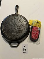 Top view of Lodge cast iron pan showing embossed USA Rushmore design on the bottom, with packaged red handle holders beside it.