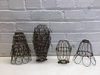 Five metal lightbulb cages of varied shapes positioned standing upright on white surface, against white brick wall, showing rust and aged wire construction.
