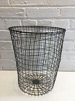 Full view of the wire mesh basket showing its cylindrical shape and black metal construction against a white brick wall.