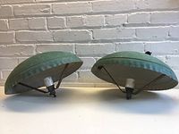 Two green metal umbrella-shaped lights sitting on a surface, showing the profile and size relative to the wall.
