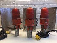 Front view of three red industrial runway lights with red textured glass globes on metal mounting plates, visible wear and attached power cables.