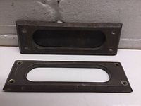 Two brass mail slot pieces; one is the outer plate with a rectangular cutout and screw holes, the other is the inner hinged flap with visible wear and patina.