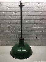 Full view of a green pendant light with a wide enamel shade and long black rod suspension against a white brick background.