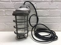 Full view of industrial cage light fixture showing the glass enclosure and metal cage with black power cord coiled beside it.