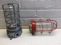 Gray industrial cage light with frosted glass globe and metal cage, sitting next to a red cage light with clear glass globe and red metal cage on a white surface against a wall.