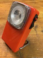 Orange rectangular vintage pocket light with circular red lens in a chrome bezel, placed upright on wooden surface.