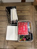 Photo showing multiple adult coloring books standing upright in a black file organizer next to an open white sketchbook and a plastic container filled with Crayola colored pencils, markers, a red spiral-bound notebook with white swirl design.