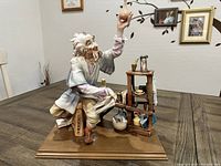 Porcelain figurine of an alchemist holding a potion flask beside a lab desk with various chemistry equipment. Figurine placed on a wooden base. Visible minor scratches on base and glued repair on desk.