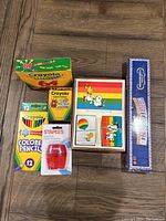 Photo of Crayola 64 crayons box, 12 colored pencils, red pencil sharpener, Snoopy Woodstock game and automatic card shuffler box on wood floor