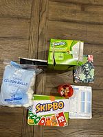 Photo showing Swiffer Sweeper dry cloth refills box, jumbo cotton balls, Skip-Bo card game box, Yahtzee dice game components, and lens care kit box on wood floor.