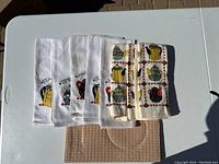 Six white kitchen towels with colorful tea pot designs and a beige textured dish drainer mat on a white table under sunlight.