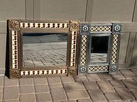 Two Southwest style metal framed mirrors: one larger brass finish with cowboy boots around the edge and one smaller pewter finish with flowers around the edge, placed on the ground in front of garage doors.