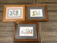 Three framed watercolor prints of whimsical dragons on wooden floor. Each print matted and framed in wood frames showing wear.