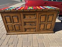 Front view of solid wood Spanish style credenza on outdoor paved ground with visible drawers and doors, color medium brown with decorative paneling and metal drawer handles, with multicolor fabric on top.