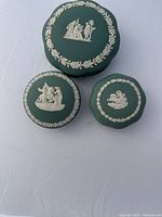Photo showing three Wedgwood jasperware boxes in teal green with white classical reliefs and wreath decorations arranged on a white cloth.