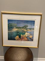 Framed art print showing the entire artwork and frame against a wall. The print depicts a coastal golf hole with rocks and trees.