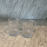 Two clear crystal beer mugs on marble surface, showing vertical etched pattern and thick crystal bases.