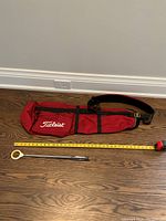 Red Titleist lightweight carry golf bag and compact telescoping ball scoop placed side by side on a wooden floor with a tape measure showing dimensions.