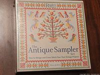 Front of unopened kit shows vintage sampler cross-stitch pattern with birds and tree design on cloth
