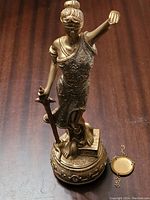 Photo of bronze-tone Lady Justice figurine front view showing detailed dress, blindfold, sword, and base with books and incomplete scales.