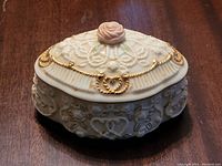 Front angled view of music box showing embossed floral and heart patterns, lace design on lid, pink rose top, and gold accents.